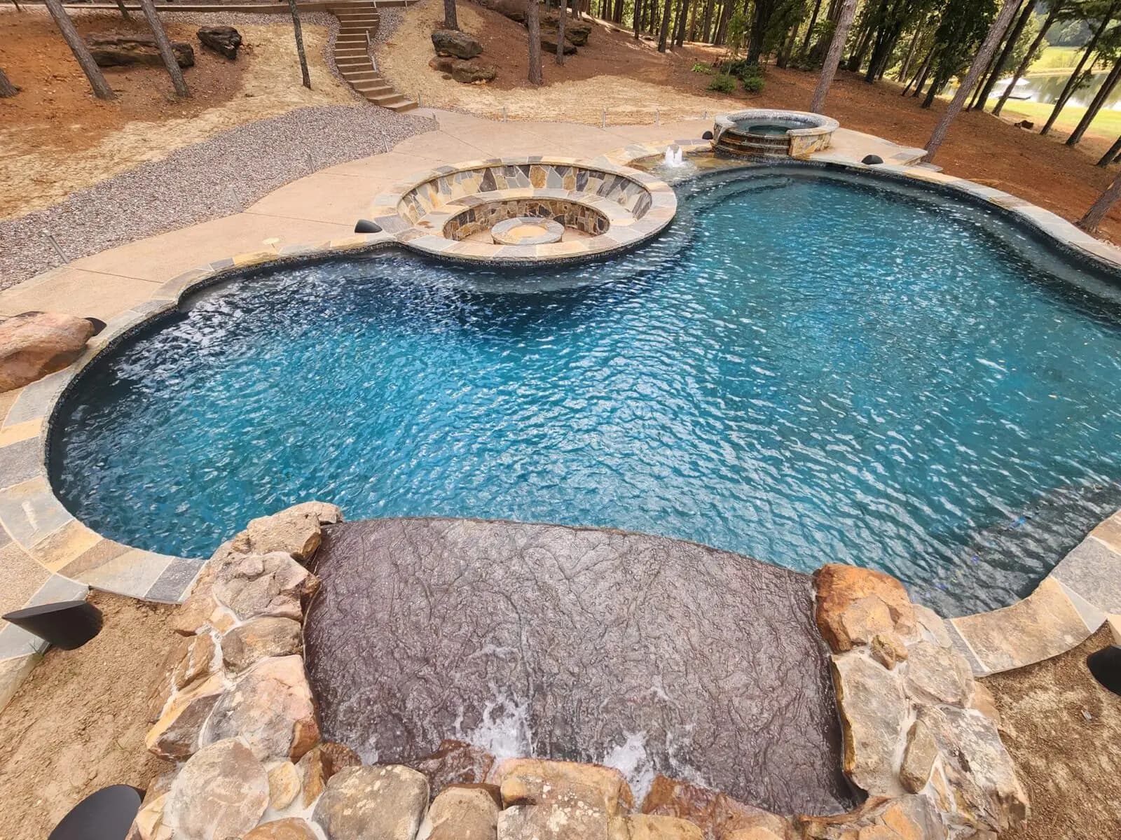 Round stone fire pit with built-in seating at the edge of a freeform pool with a rock waterfall and a spa