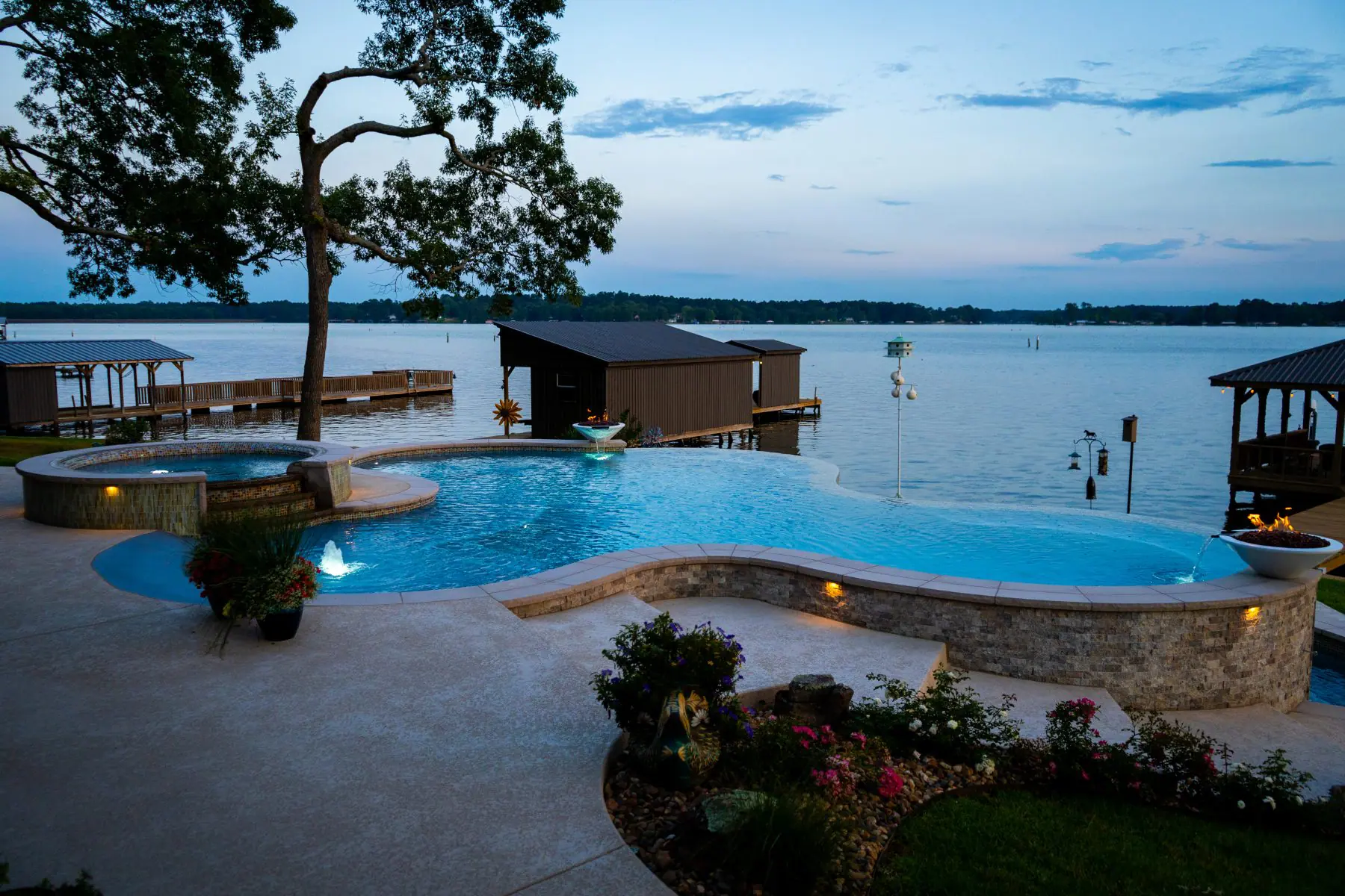 Custom freeform gunite pool with a raised spa and landscaped patio beside a lake at dusk