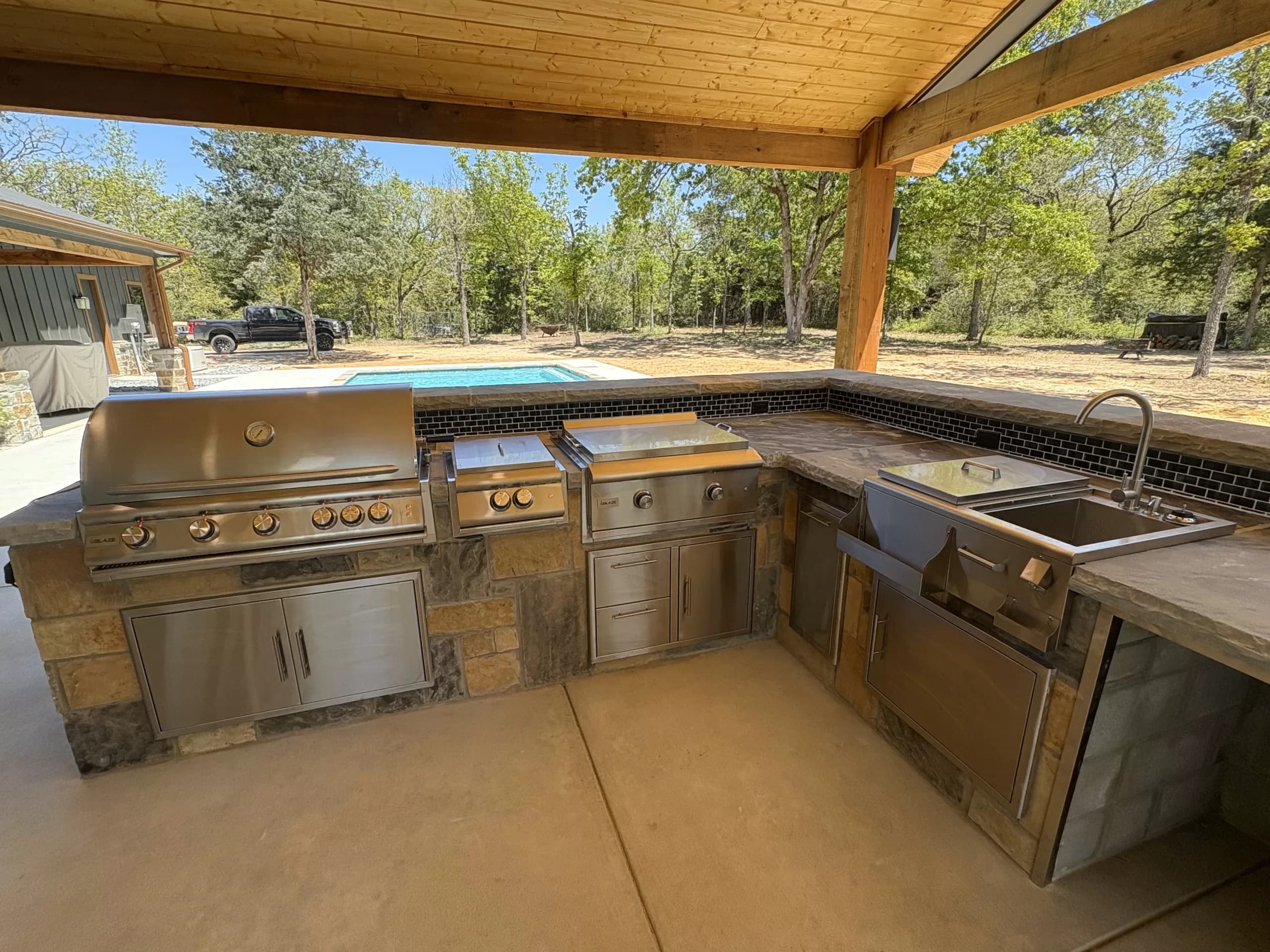 Outdoor kitchen with a stainless steel grill, a stone counter and a pool in the background