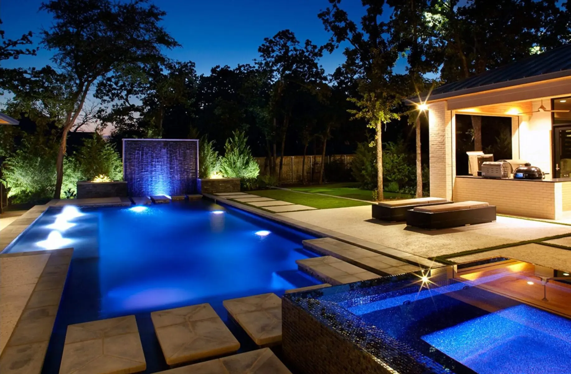 Lit custom pool at dusk with a water wall, a raised spa and a covered outdoor kitchen