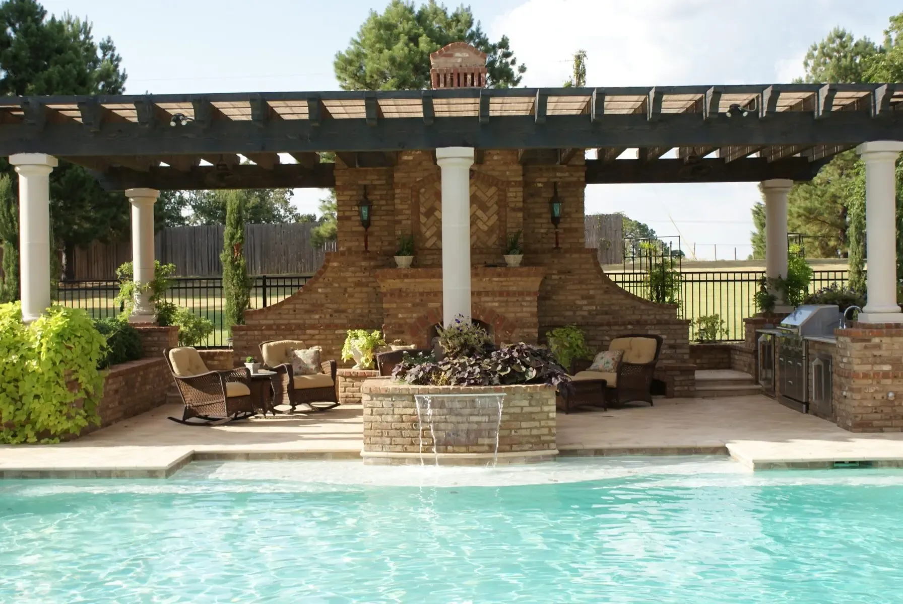 Covered pergola with a brick fireplace, seating and an outdoor kitchen beside a pool with a waterfall feature