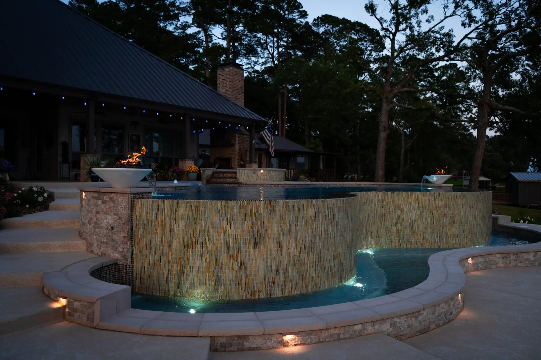 Infinity edge pool with a glass tile wall and fire bowls at dusk